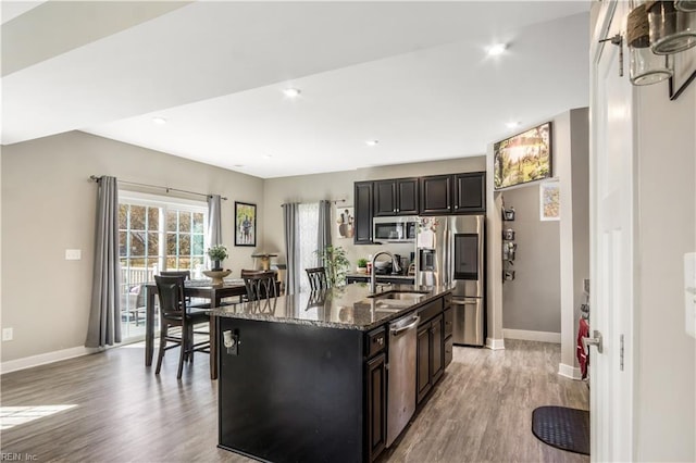 kitchen featuring appliances with stainless steel finishes, a kitchen island with sink, a sink, dark stone countertops, and light wood-type flooring