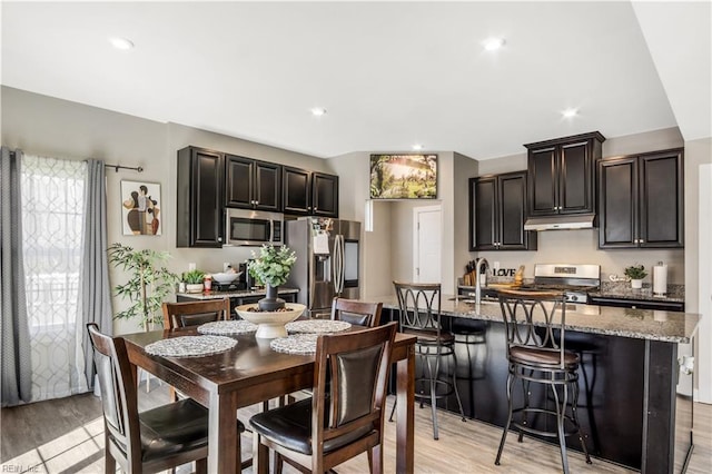 kitchen with light stone counters, stainless steel appliances, light wood-style flooring, an island with sink, and under cabinet range hood