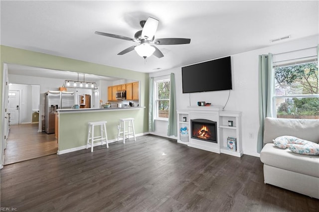 living area with a warm lit fireplace, dark wood-type flooring, visible vents, baseboards, and a ceiling fan