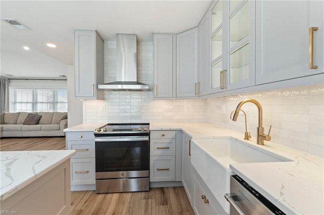 kitchen featuring visible vents, glass insert cabinets, stainless steel appliances, light wood-type flooring, and wall chimney range hood