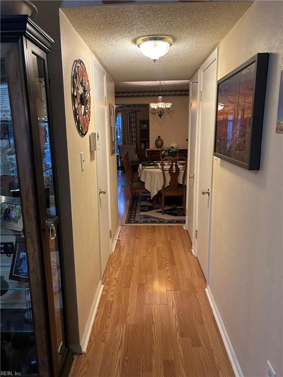 corridor with light wood-type flooring, an inviting chandelier, baseboards, and a textured ceiling