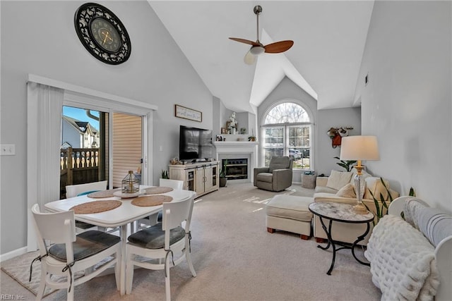 dining area with ceiling fan, high vaulted ceiling, light colored carpet, baseboards, and a glass covered fireplace