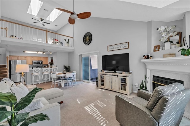 living room featuring light carpet, a skylight, a glass covered fireplace, ceiling fan, and high vaulted ceiling