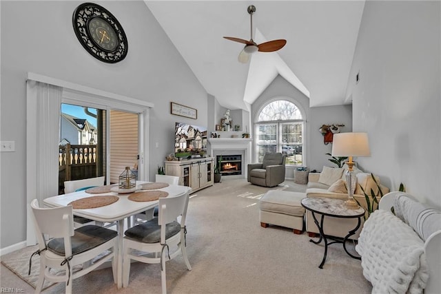 dining space featuring light carpet, ceiling fan, high vaulted ceiling, a warm lit fireplace, and baseboards