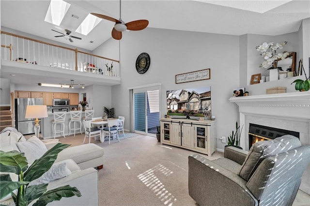 living room featuring light carpet, a skylight, a glass covered fireplace, ceiling fan, and high vaulted ceiling