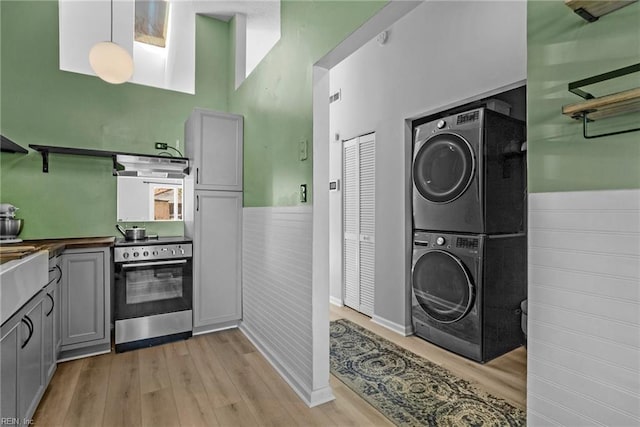 kitchen with gray cabinetry, stacked washer / drying machine, light wood-type flooring, wainscoting, and stainless steel electric stove