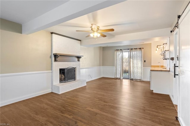 unfurnished living room with a wainscoted wall, a barn door, a brick fireplace, and dark wood-style flooring