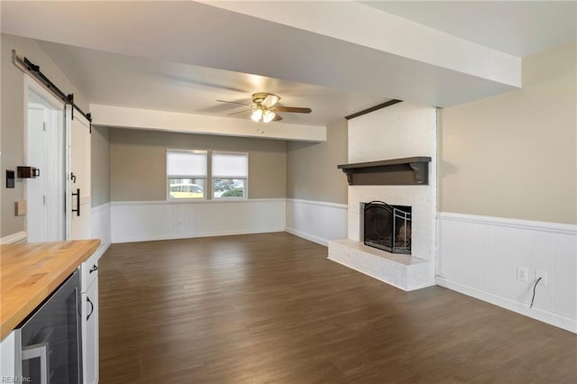 unfurnished living room featuring a barn door, wine cooler, a wainscoted wall, a fireplace, and a ceiling fan