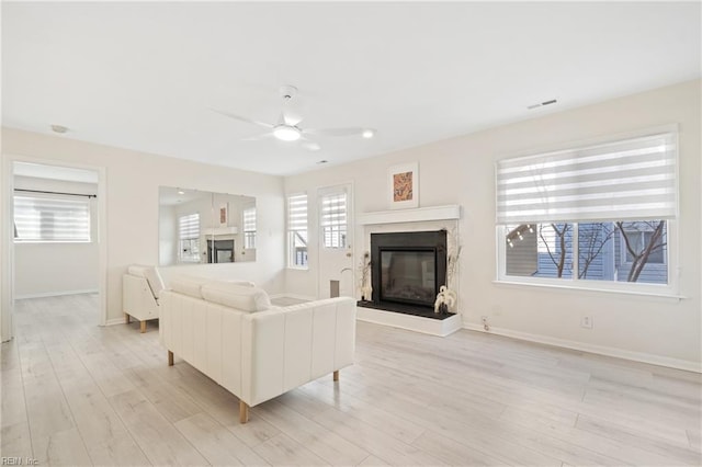 living room with ceiling fan, light wood-style flooring, visible vents, baseboards, and a glass covered fireplace