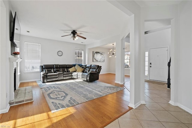 living room with ceiling fan with notable chandelier, a fireplace with raised hearth, baseboards, and light tile patterned floors