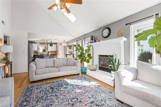 living room featuring vaulted ceiling with skylight, visible vents, a glass covered fireplace, light wood-style flooring, and ceiling fan