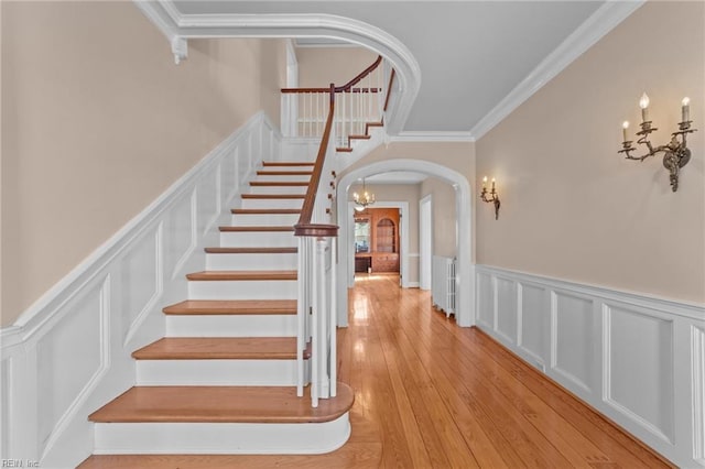 staircase featuring arched walkways, a decorative wall, wainscoting, hardwood / wood-style floors, and crown molding