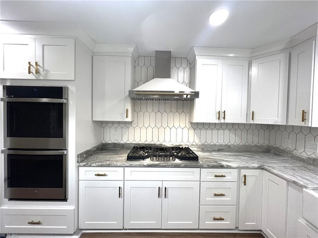 kitchen featuring light stone counters, stainless steel appliances, white cabinets, backsplash, and wall chimney exhaust hood