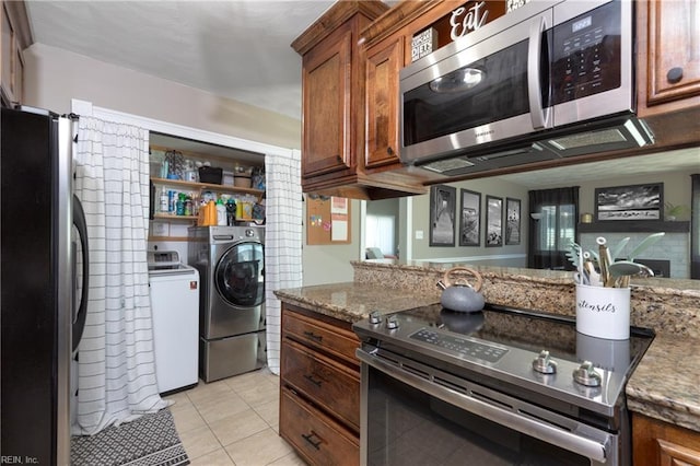 kitchen with brown cabinetry, light stone countertops, stainless steel appliances, washing machine and dryer, and light tile patterned flooring