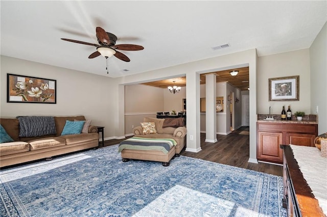 living room with dark wood-style flooring, visible vents, baseboards, and ceiling fan with notable chandelier