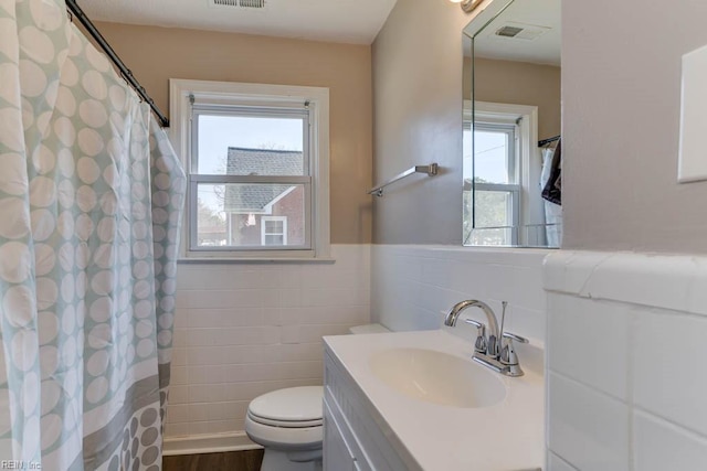 bathroom with toilet, visible vents, vanity, tile walls, and wainscoting