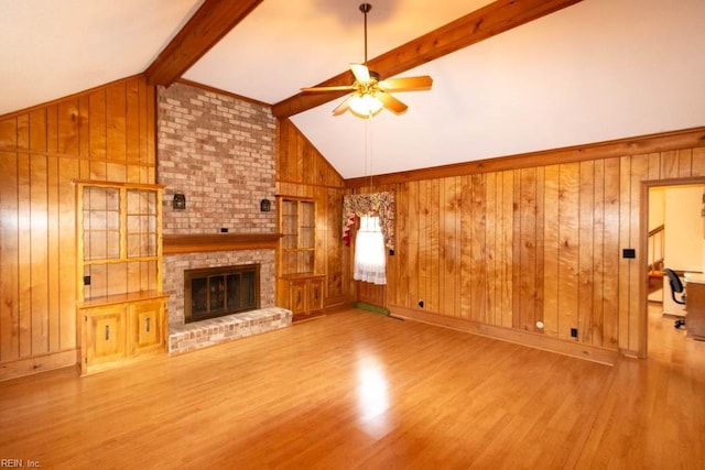 unfurnished living room with wooden walls, lofted ceiling with beams, ceiling fan, wood finished floors, and a brick fireplace