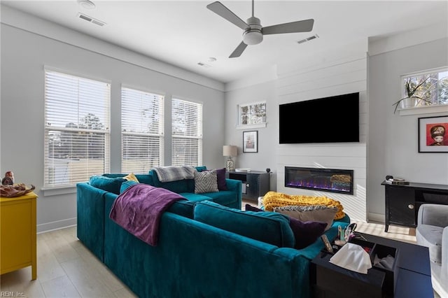 living room with ceiling fan, light wood-type flooring, a glass covered fireplace, and visible vents