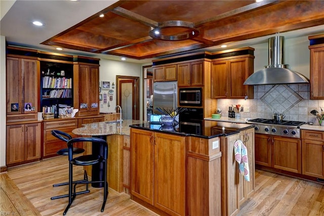 kitchen with stainless steel appliances, an island with sink, wall chimney exhaust hood, and light wood finished floors