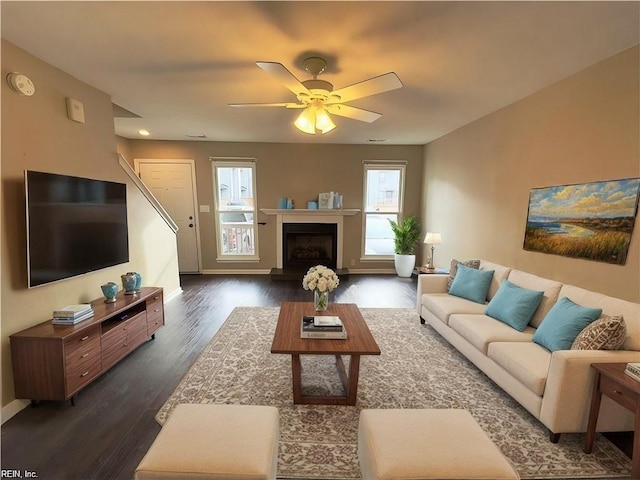 living room with ceiling fan, a fireplace with raised hearth, dark wood-type flooring, and baseboards