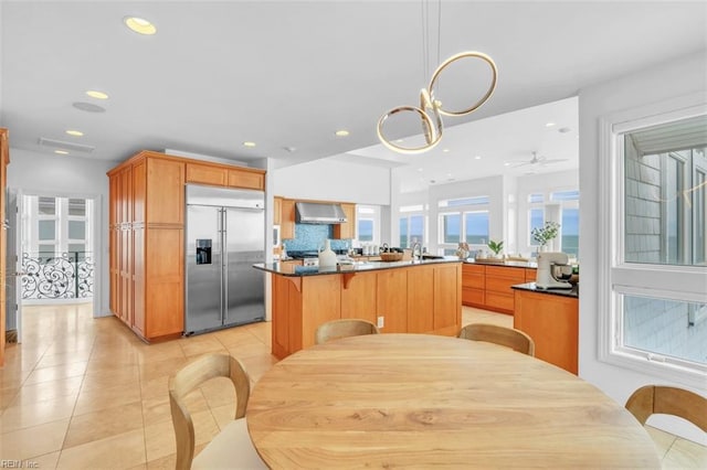 kitchen with light tile patterned floors, dark countertops, stainless steel built in fridge, wall chimney range hood, and a sink