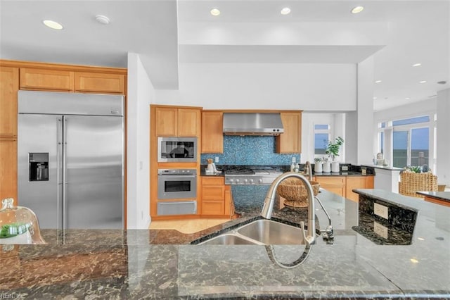 kitchen with decorative backsplash, a sink, dark stone countertops, built in appliances, and wall chimney exhaust hood