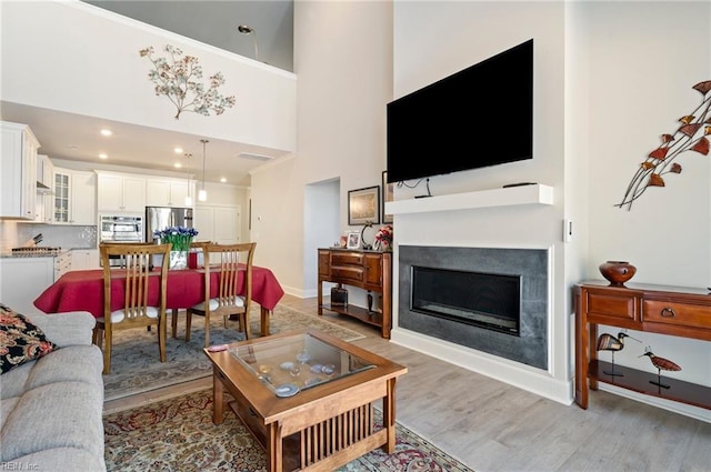 living room with recessed lighting, a towering ceiling, light wood-style flooring, a glass covered fireplace, and baseboards