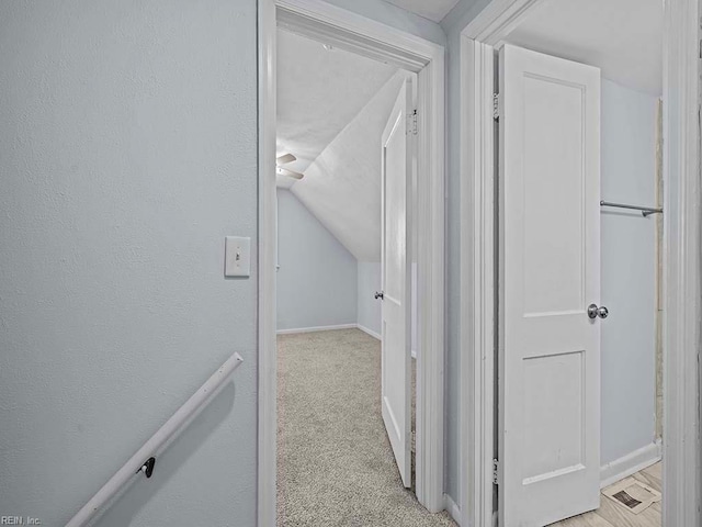 corridor with lofted ceiling, a textured wall, baseboards, and light colored carpet