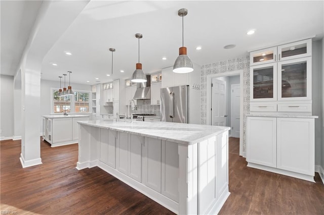 kitchen featuring white cabinets, dark wood finished floors, stainless steel refrigerator, a large island with sink, and wall chimney range hood