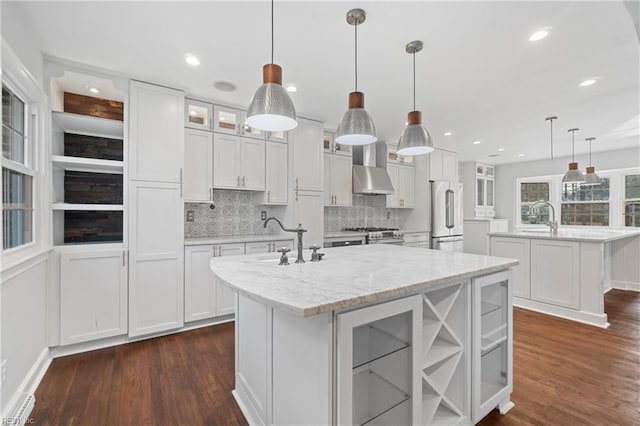kitchen with high end refrigerator, white cabinets, wall chimney exhaust hood, a kitchen island with sink, and a sink