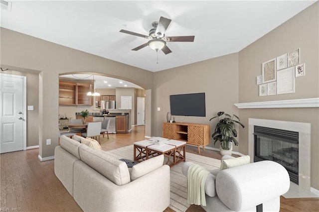 living room featuring arched walkways, light wood-style flooring, a ceiling fan, baseboards, and a glass covered fireplace