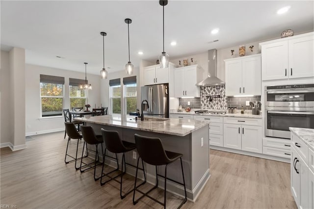 kitchen featuring tasteful backsplash, appliances with stainless steel finishes, light wood-style floors, an island with sink, and wall chimney exhaust hood