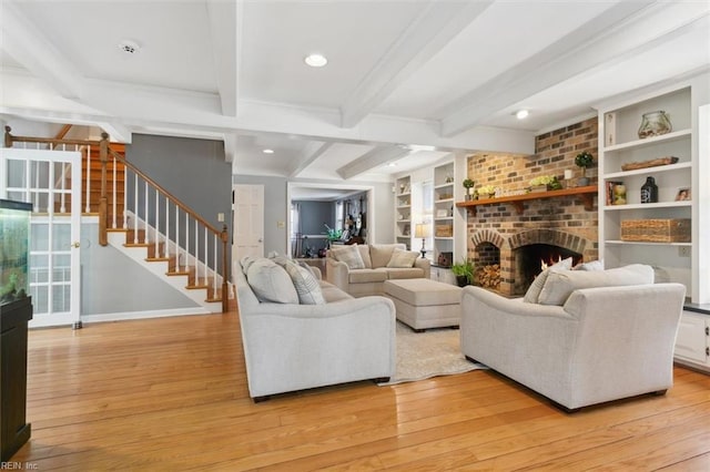 living room with built in shelves, stairs, light wood-type flooring, a brick fireplace, and beamed ceiling