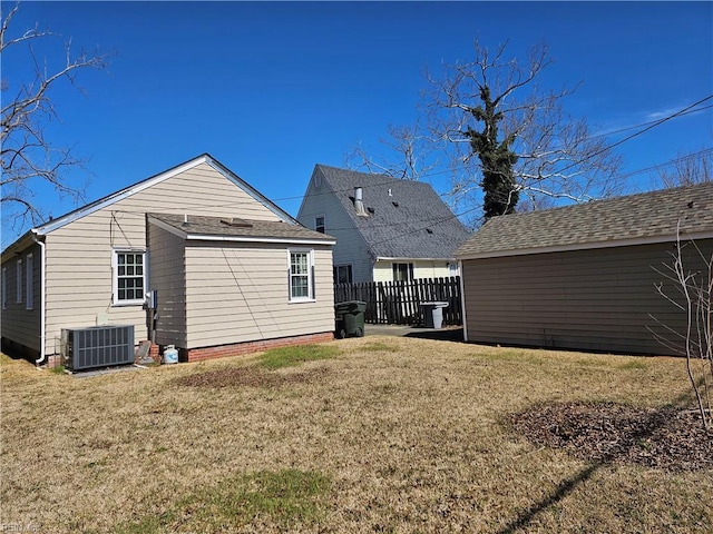 rear view of property featuring fence, central AC unit, a lawn, and roof with shingles