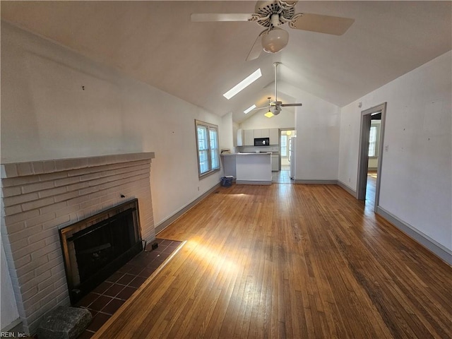 unfurnished living room with wood-type flooring, a brick fireplace, ceiling fan, vaulted ceiling with skylight, and baseboards