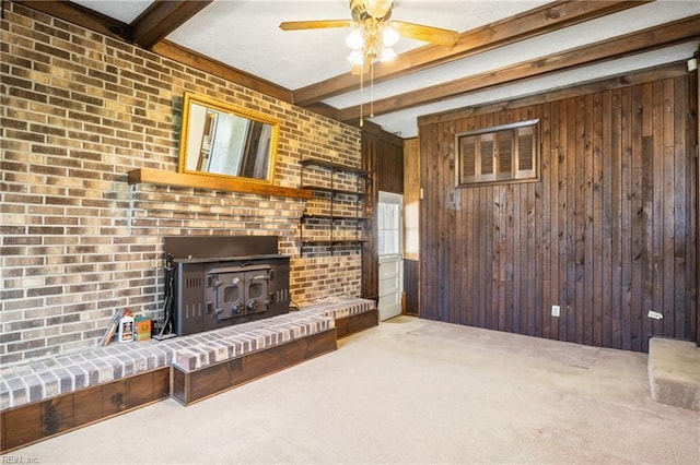 carpeted living area with beamed ceiling, wood walls, a wood stove, and a ceiling fan