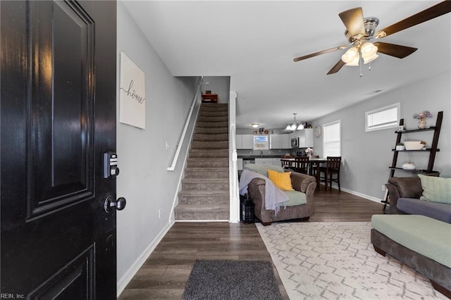 living area featuring stairs, dark wood-type flooring, ceiling fan with notable chandelier, and baseboards