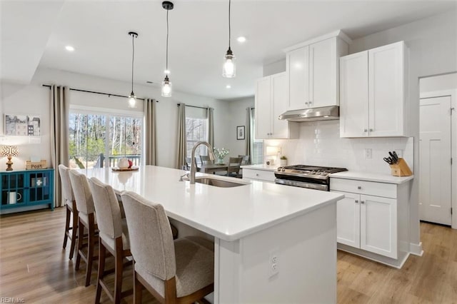 kitchen featuring tasteful backsplash, an island with sink, a sink, under cabinet range hood, and stainless steel range with gas stovetop