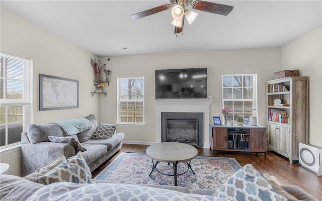 living area with dark wood-style floors, a glass covered fireplace, a wealth of natural light, and a ceiling fan