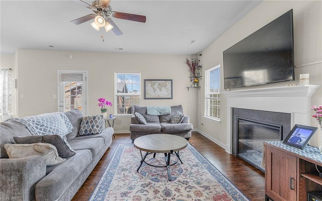 living room featuring ceiling fan, dark wood-type flooring, a glass covered fireplace, and baseboards