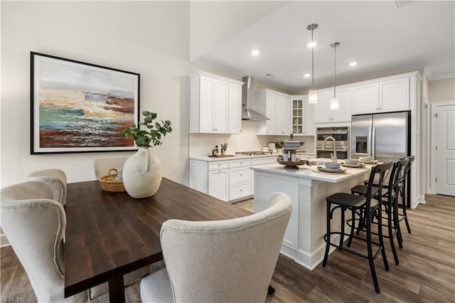 kitchen featuring a center island with sink, wall chimney exhaust hood, glass insert cabinets, wood finished floors, and stainless steel appliances