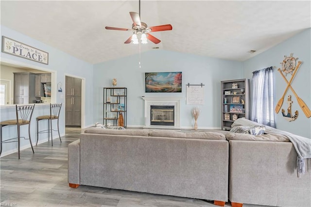 living area featuring a glass covered fireplace, vaulted ceiling, ceiling fan, light wood-type flooring, and baseboards