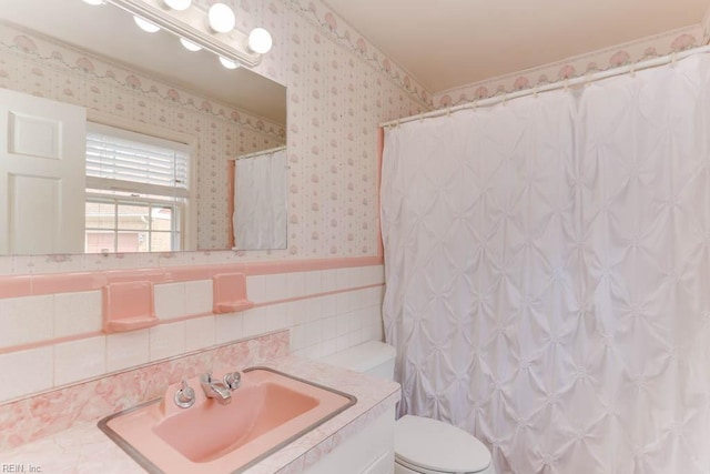 bathroom featuring wallpapered walls, wainscoting, toilet, vanity, and tile walls