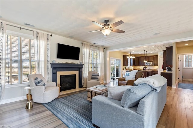 living room with visible vents, light wood-style floors, a glass covered fireplace, baseboards, and ceiling fan with notable chandelier