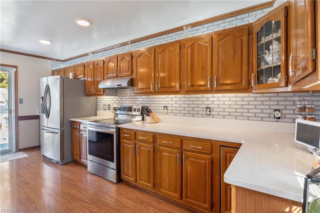 kitchen featuring brown cabinets, crown molding, appliances with stainless steel finishes, glass insert cabinets, and under cabinet range hood