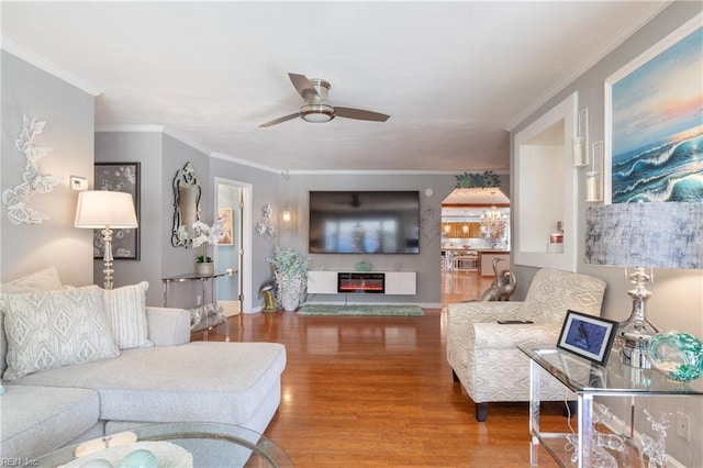 living room featuring arched walkways, a glass covered fireplace, ornamental molding, wood finished floors, and ceiling fan with notable chandelier