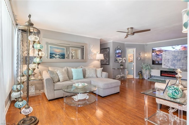 living area with wood finished floors, a ceiling fan, baseboards, ornamental molding, and a glass covered fireplace