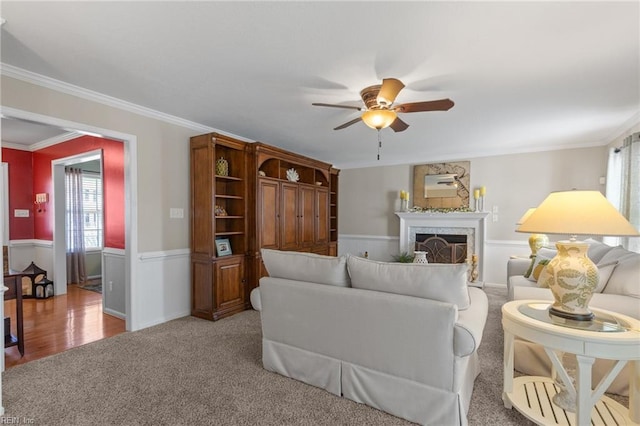 living room featuring light colored carpet, a wainscoted wall, ceiling fan, crown molding, and a high end fireplace