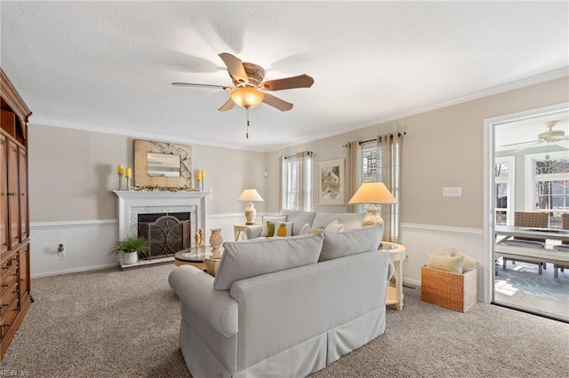 carpeted living room featuring a fireplace, ornamental molding, a ceiling fan, and wainscoting