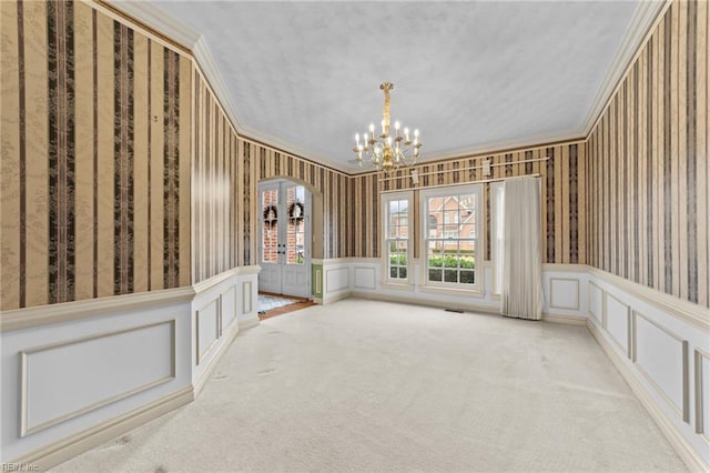 carpeted empty room featuring crown molding, a decorative wall, wainscoting, a chandelier, and wallpapered walls